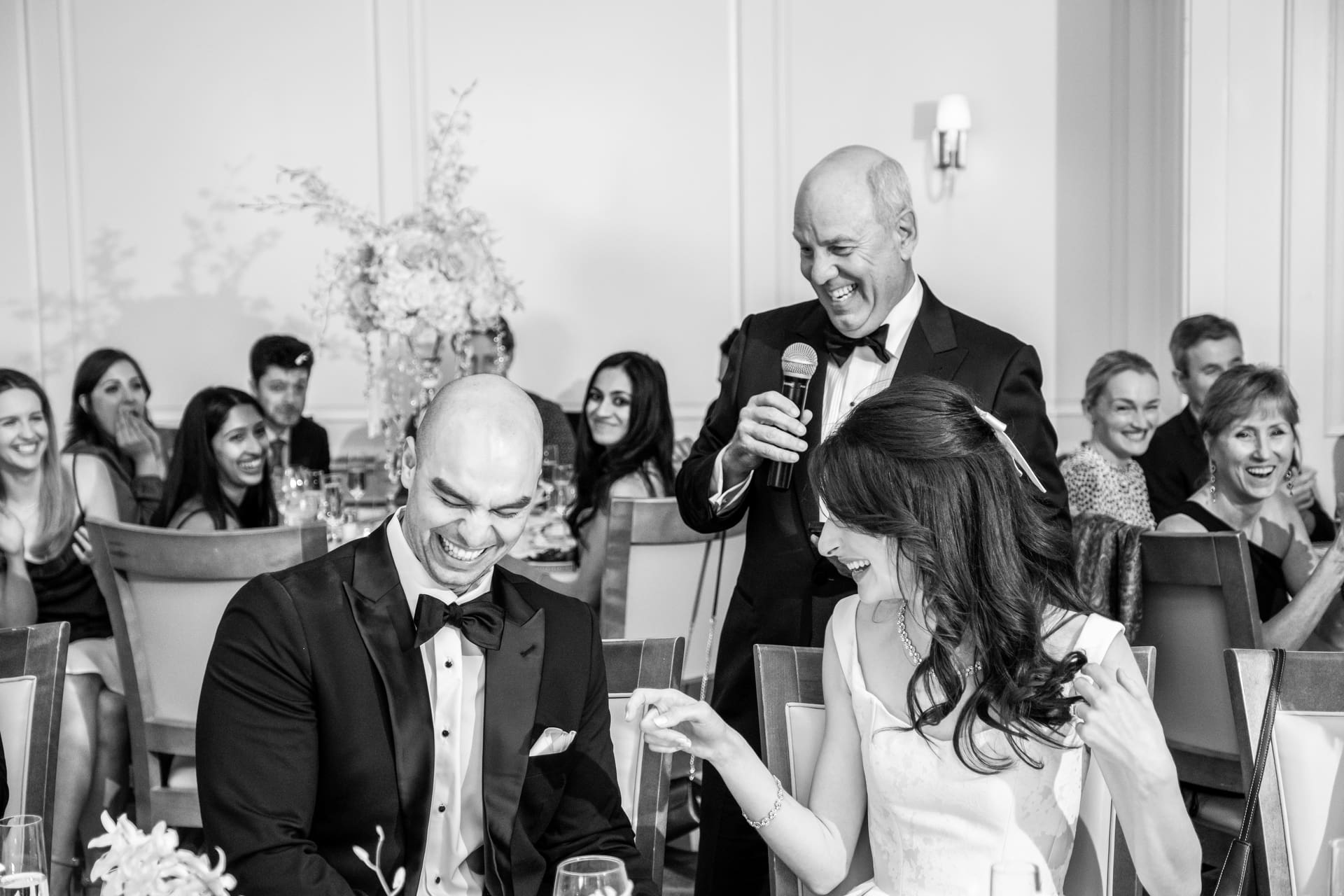 Groom laughing as a wedding guest gives a speech while the bride and guests react