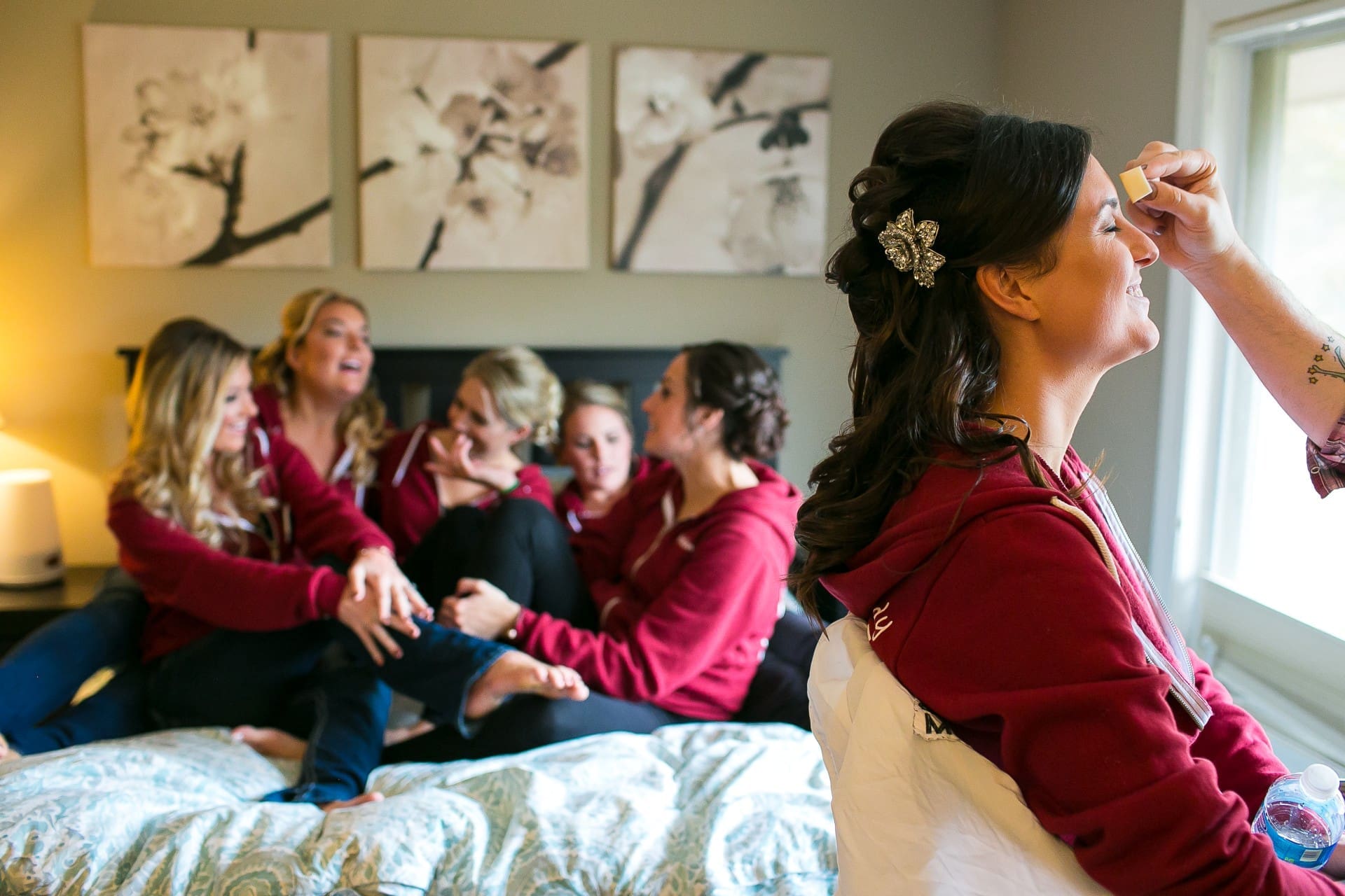 Bride having her makeup applied while her bridesmaids share a joyful moment before the wedding ceremony.