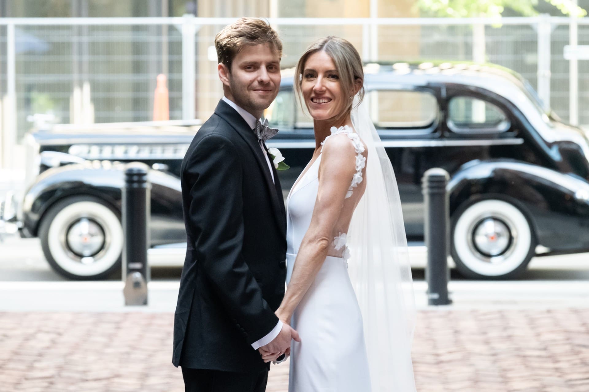 Newlywed couple holding hands and smiling beside a vintage car.