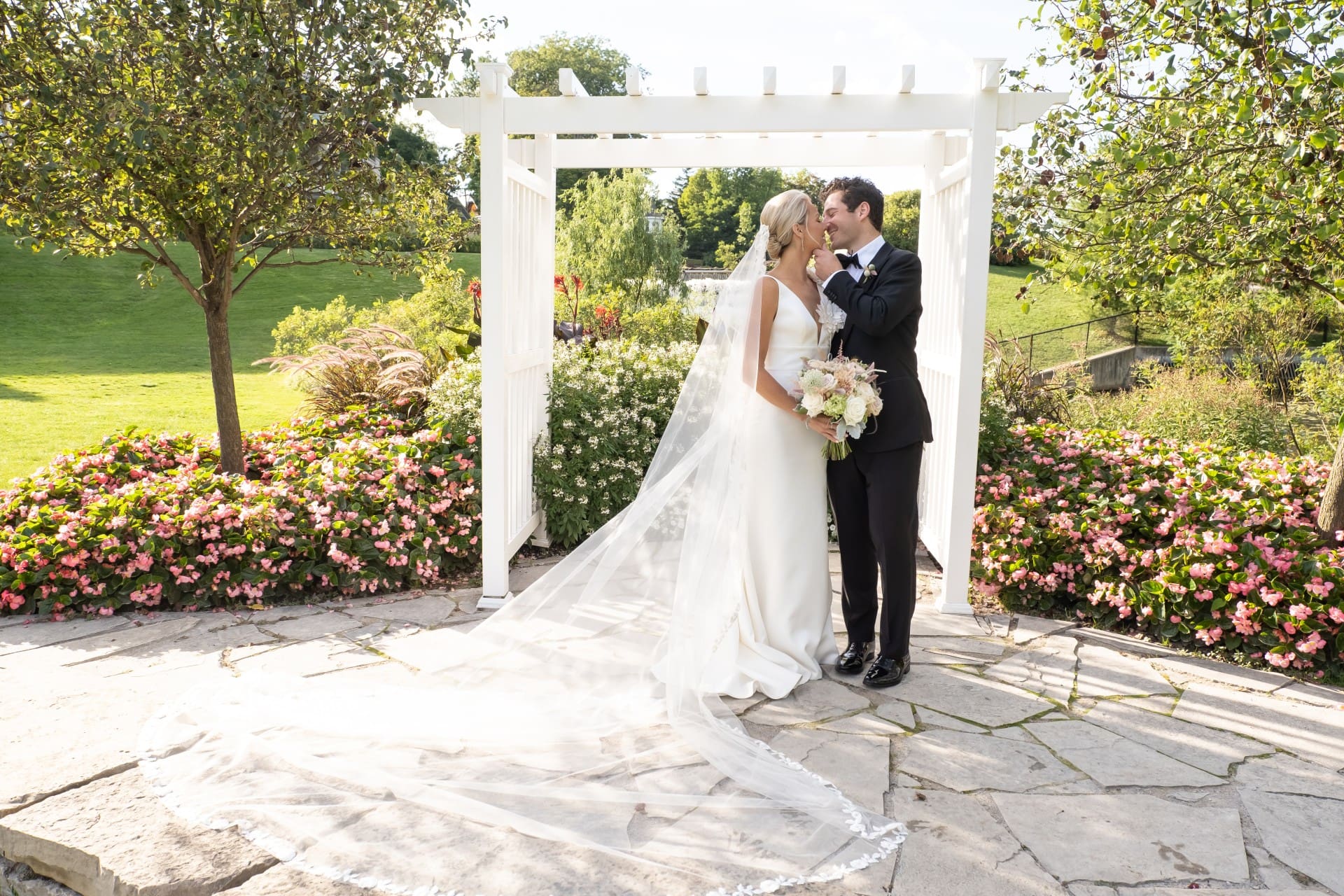Bride and groom embracing beneath a white garden arch surrounded by flowers and greenery