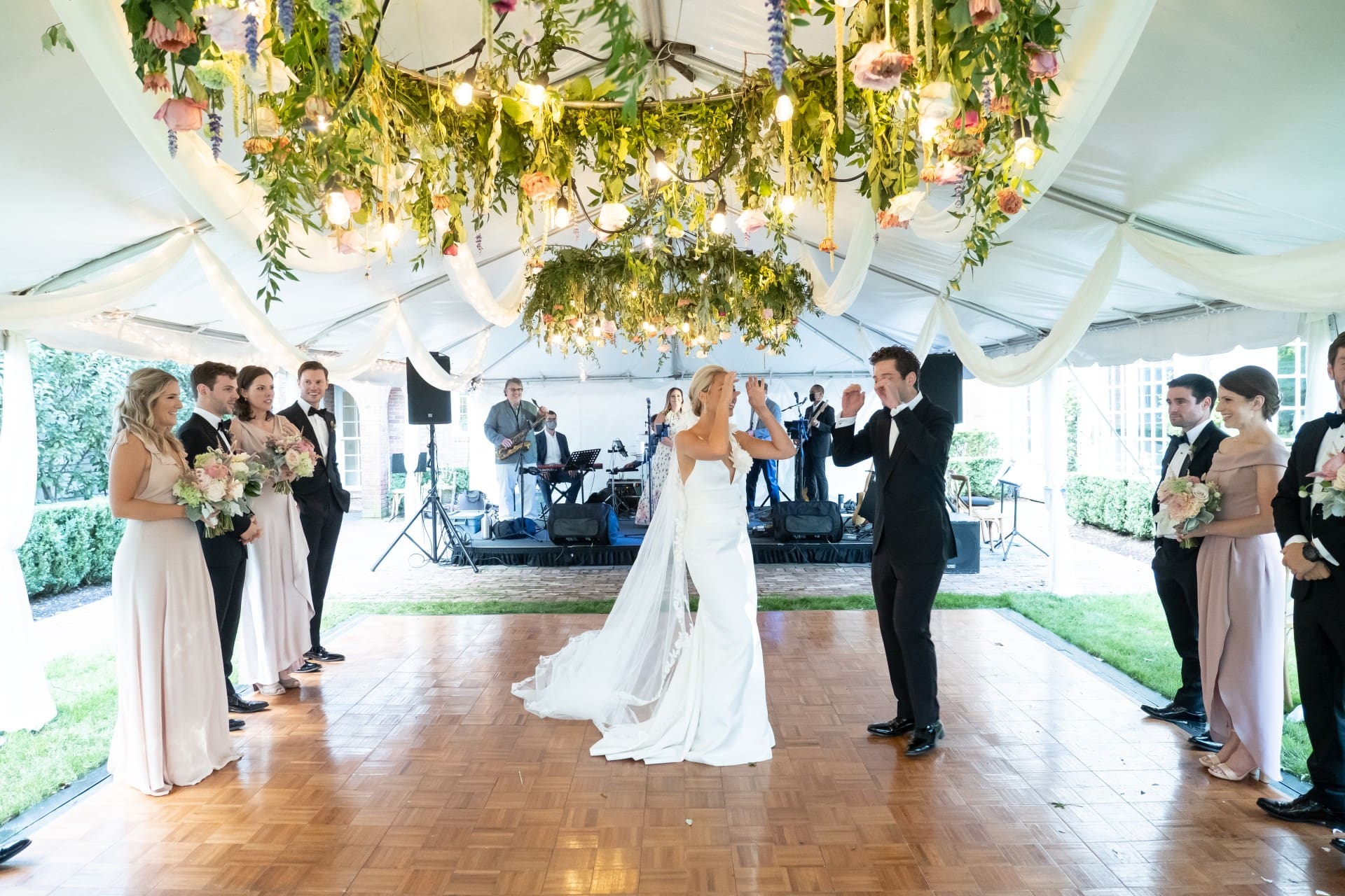 Bride and groom dancing beneath a floral reception canopy with their wedding party nearby